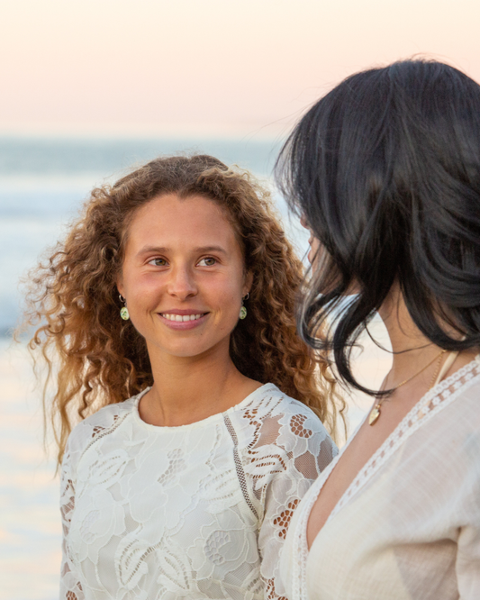 Two women standing on a beach at sunset, with the woman on the left wearing a white lace top and looking toward the camera, while the woman on the right is shown from the back wearing a cream lace top.